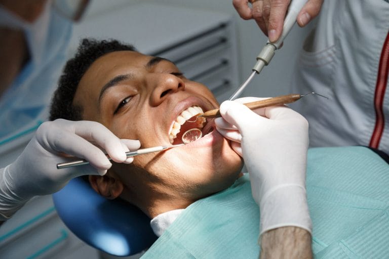 Dentist examining oral cavity of young African-American man working in in dental clinic with assistant